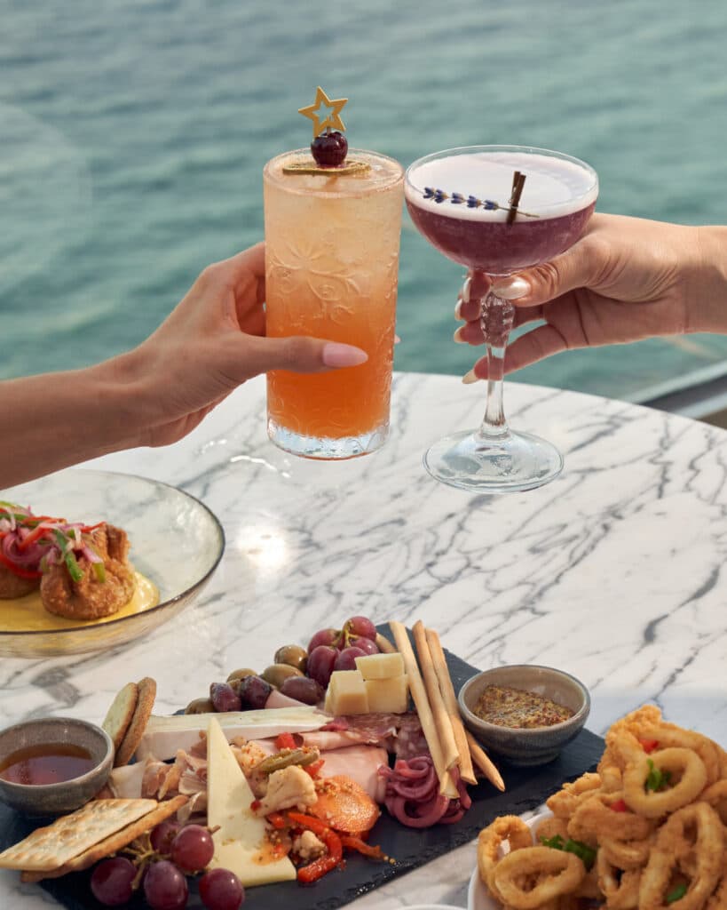 two people cheersing cocktails over charcuterie and food next to the ocean